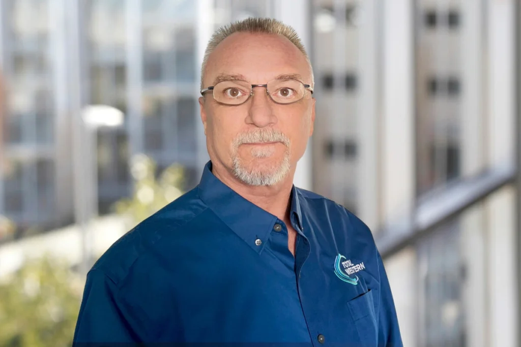 Corporate portrait of an employee wearing a blue button-down shirt with the Total Western logo, standing in front of a modern urban building backdrop.