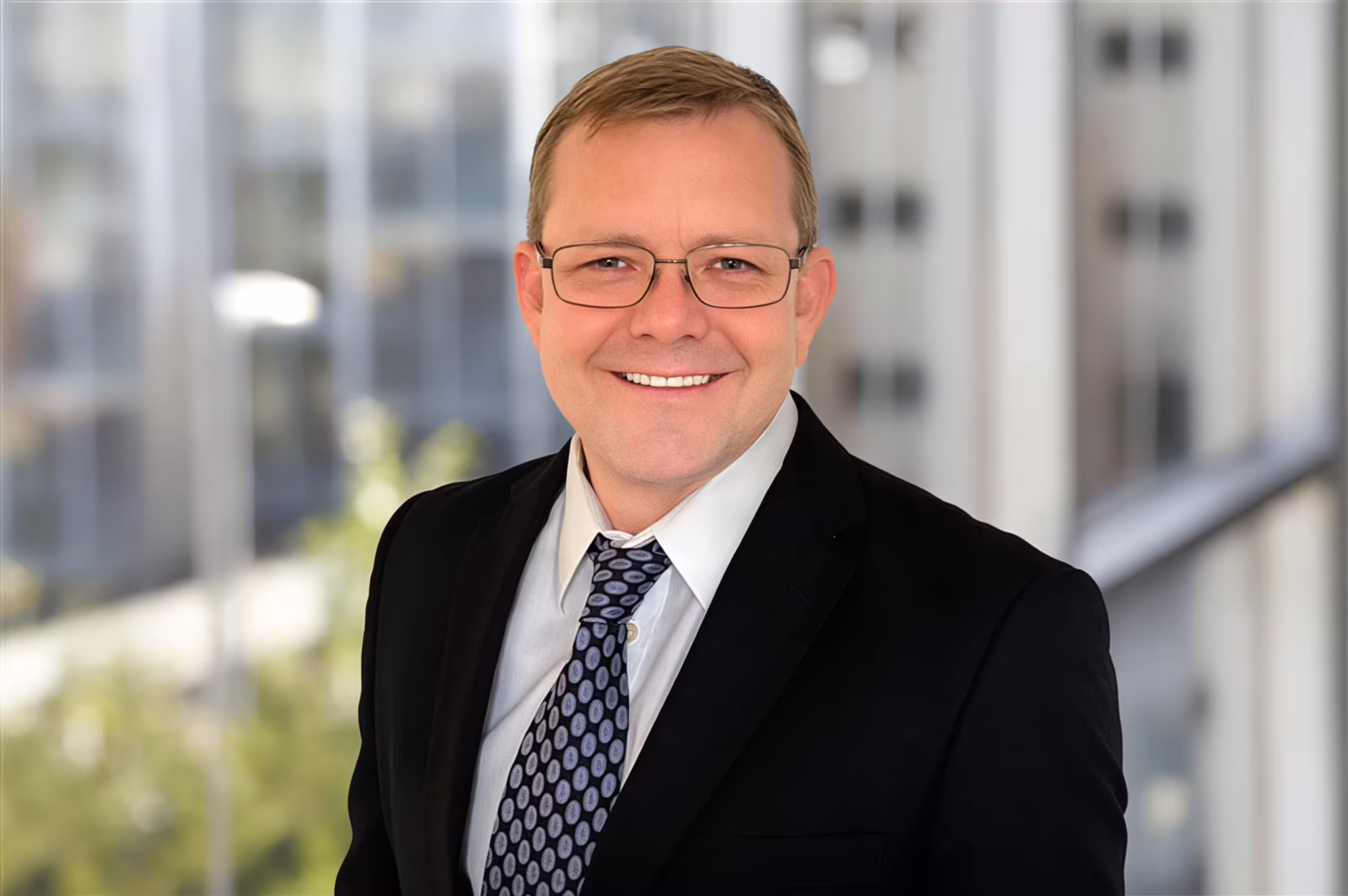Professional business portrait of a smiling man in a black suit and patterned tie, wearing glasses, standing in a modern office with cityscape background.